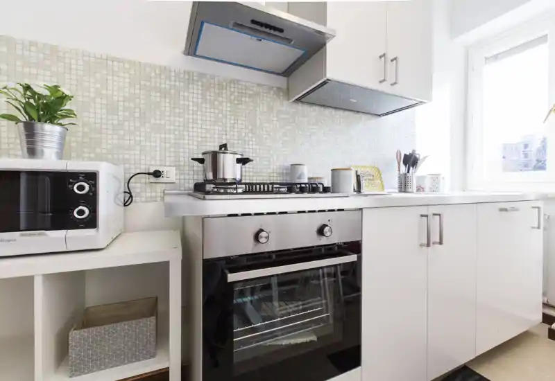 Bright, modern kitchen with a built-in oven, gas hob, microwave and countertop storage; natural light from the window highlights the clean white cabinets and tiled backsplash.