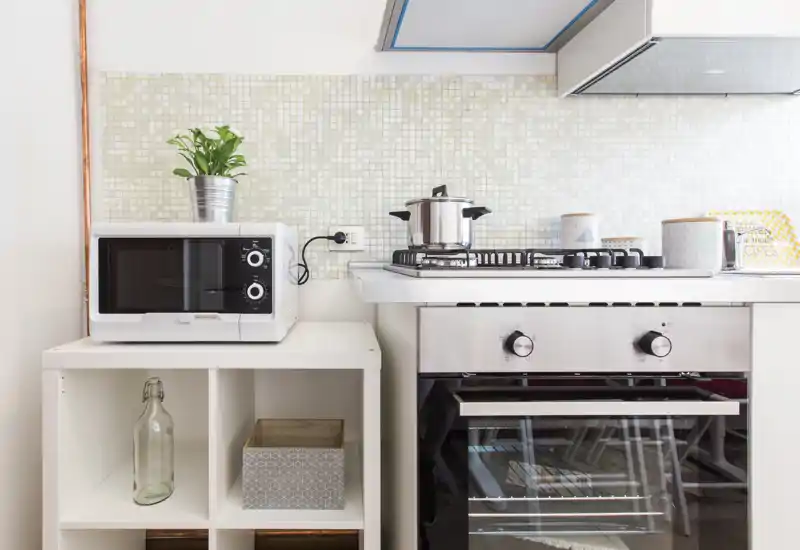 Bright, modern kitchen corner with a gas hob and oven, microwave on a shelf, and small decorative touches like a potted plant and storage boxes.