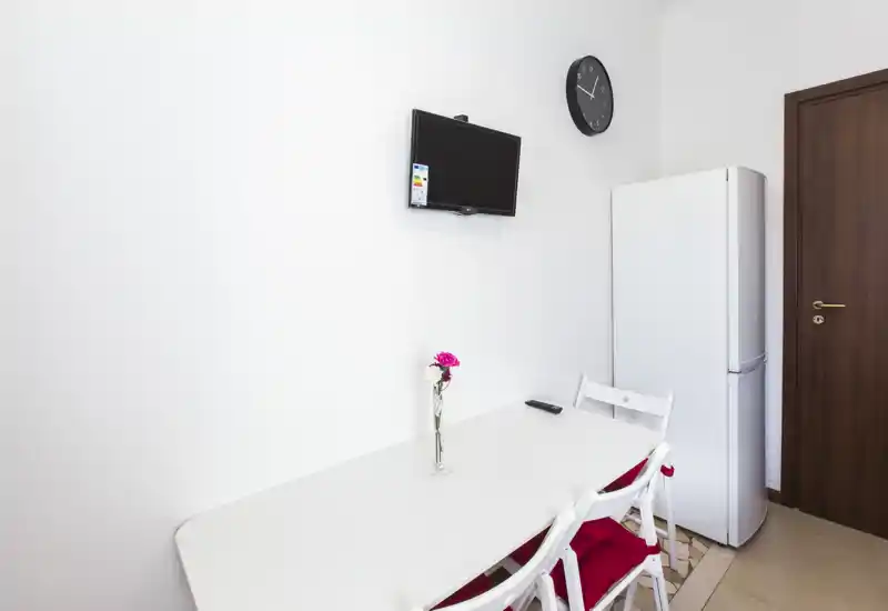 Compact kitchen/dining corner with a white table, two chairs with red cushions, a tall fridge and wall-mounted TV; bright, minimal and neatly arranged.