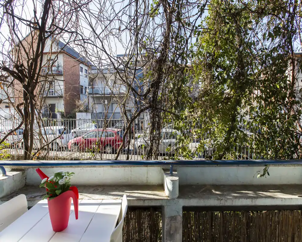 Piccolo balcone soleggiato con un tavolo bianco e un annaffiatoio-vaso rosso, che si affaccia su una strada parzialmente schermata da rampicanti.