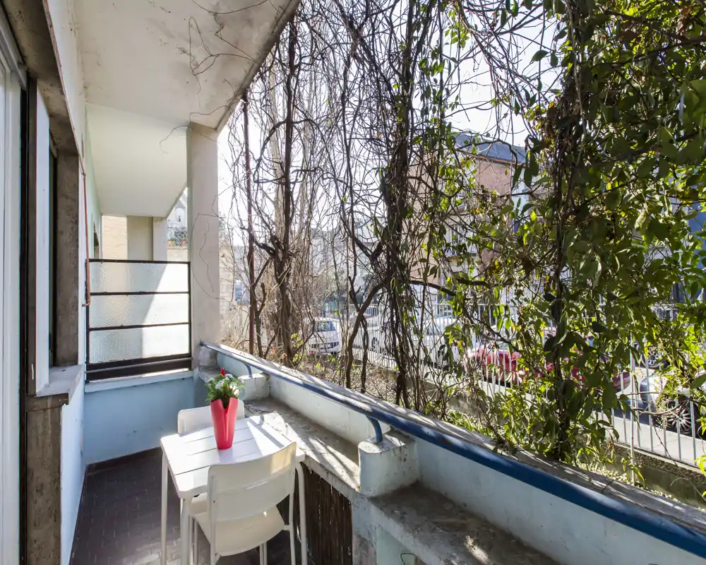 Balcone soleggiato con un piccolo tavolo e due sedie, affacciato su una strada alberata — perfetto per il caffè mattutino.