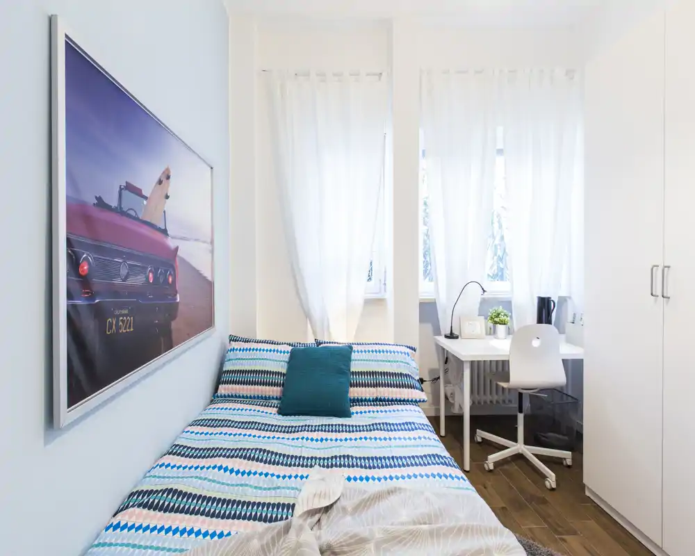 Bright, compact bedroom with a double bed dressed in patterned blue bedding, a teal accent cushion, white wardrobe and a small desk by the window letting in soft natural light.