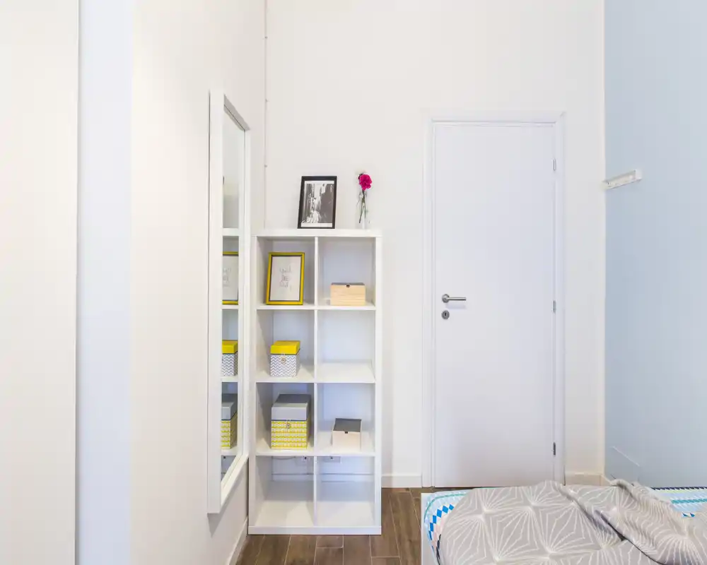 Bright, minimal bedroom corner with a glimpse of the bed, a white cubby shelving unit with decorative storage boxes, a full-length mirror and a closed door — modern, tidy and well lit.