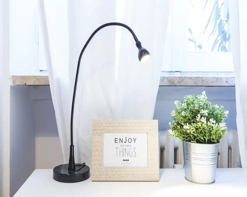 Bright, minimal decorative tabletop with a flexible reading lamp, framed print that says 'Enjoy the little things' and a small potted plant on a white surface beneath a window.