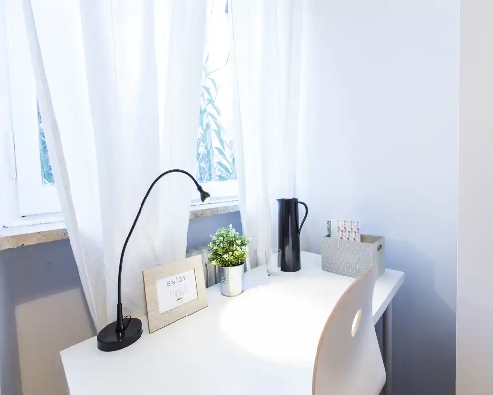 A bright, minimal bedside or desk area with white curtains, a small plant and a lamp — suitable for a calm bedroom workspace.