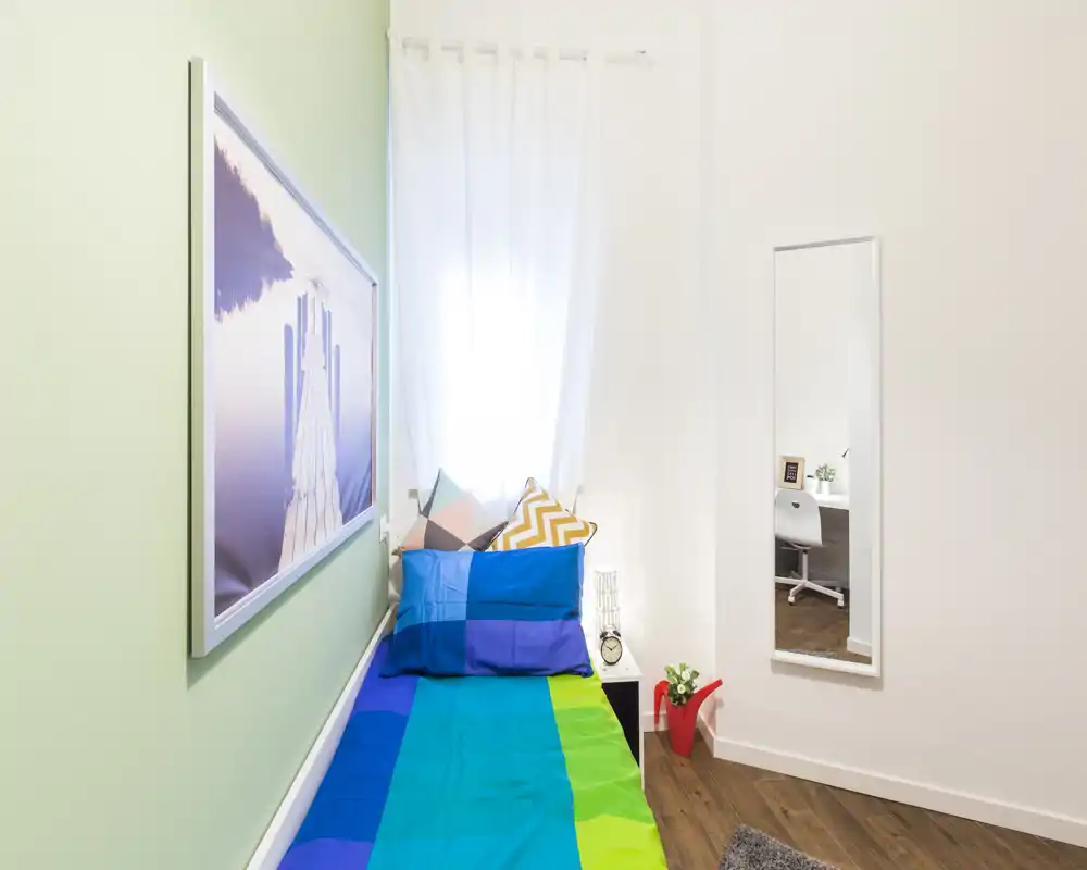 Camera luminosa e compatta con un letto singolo colorato, cuscini decorativi e una finestra che diffonde luce naturale morbida — perfetta per un soggiorno accogliente da solo.