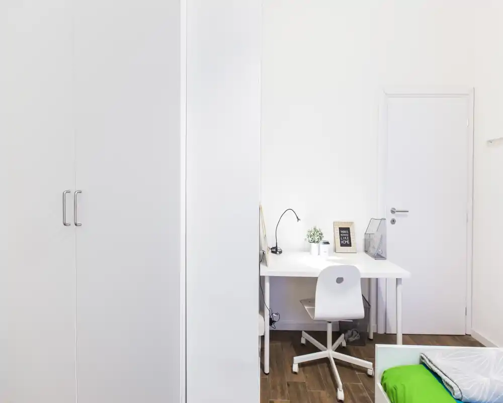 Angolo camera da letto luminoso e compatto con scrivania e letto parzialmente visibile con biancheria verde. Mobili bianchi e pavimento in legno dal design semplice e moderno.