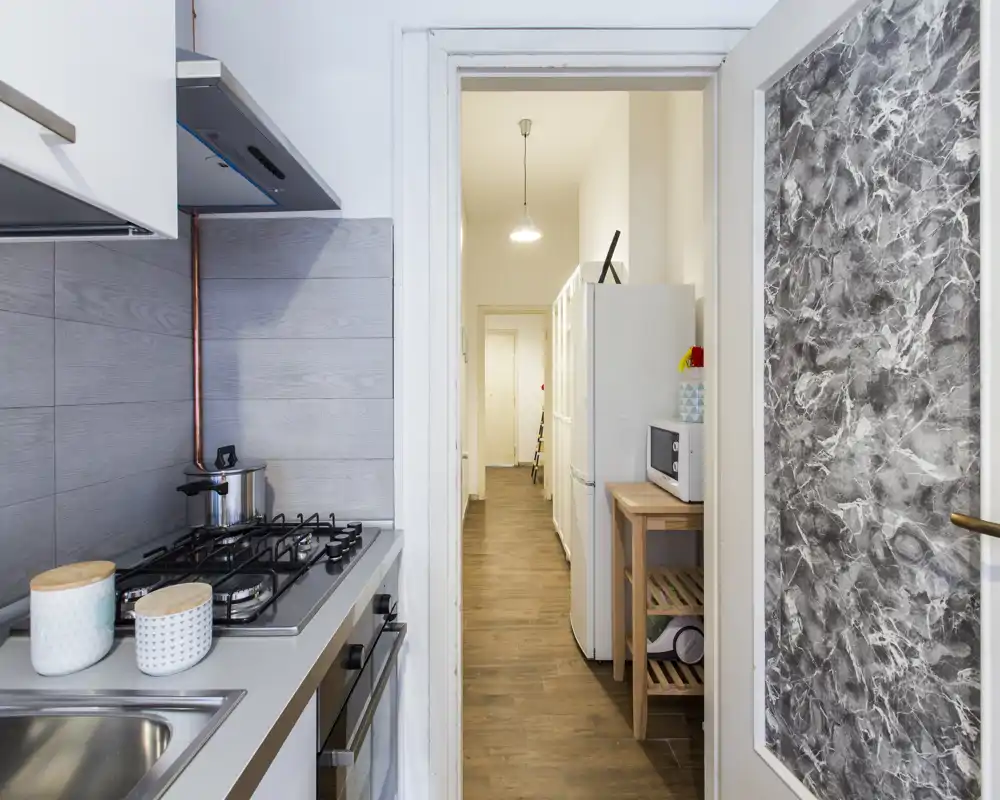 Compact modern kitchen with gas hob, stainless sink and tiled splashback; neat counter with canisters and a view down a wooden-floored corridor.