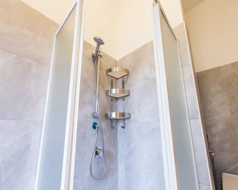 Modern corner shower with frosted sliding doors, handheld shower head and stainless steel corner shelves; clean light-gray tile walls create a contemporary, easy-to-maintain bathroom space.