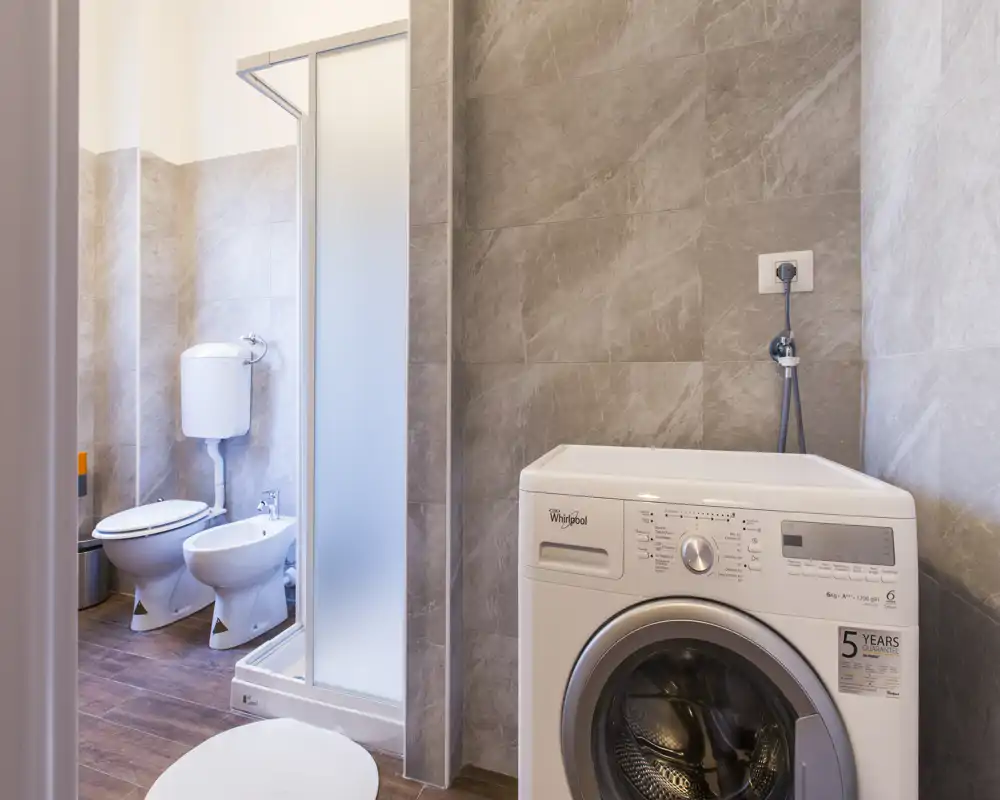 Bright, modern bathroom with a shower cabin, toilet, bidet and a front-loading washing machine against textured stone-effect tiles.