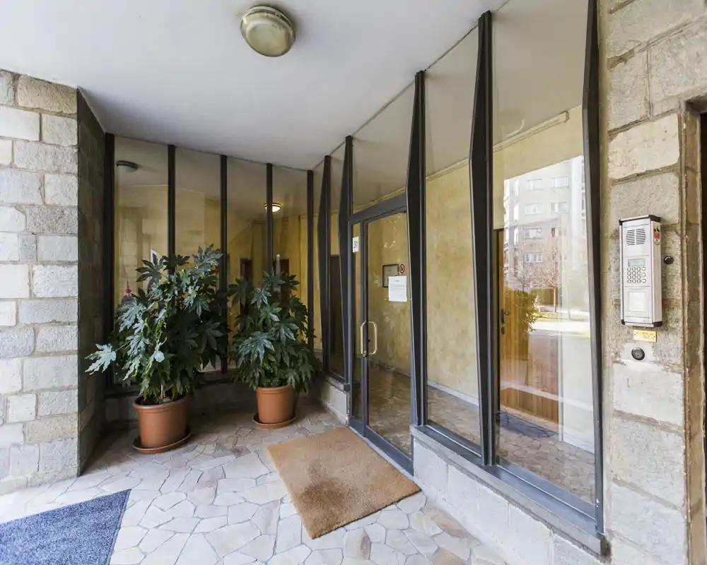 Well-lit residential entrance with glass doors, intercom panel and decorative potted plants, offering a clean and welcoming lobby area.