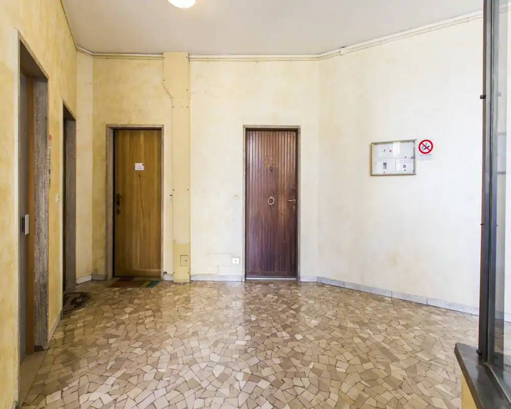 Spacious building entrance/hall with tiled mosaic floor, two wooden doors and neutral painted walls — bright and uncluttered common area ideal for apartment access.