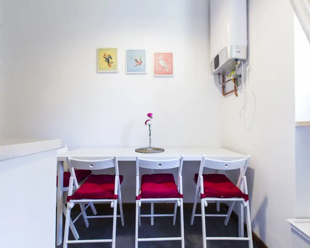 Compact dining nook with a white table and four folding chairs accented by red cushions; bright, minimal and neatly styled with a single flower centerpiece.