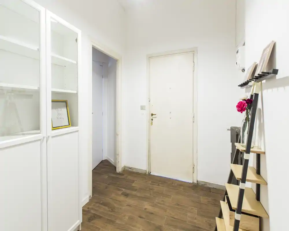 Bright, minimal entrance hallway with wood-effect flooring, a white main door and simple shelving for shoes or décor; clean and functional first-impression space.