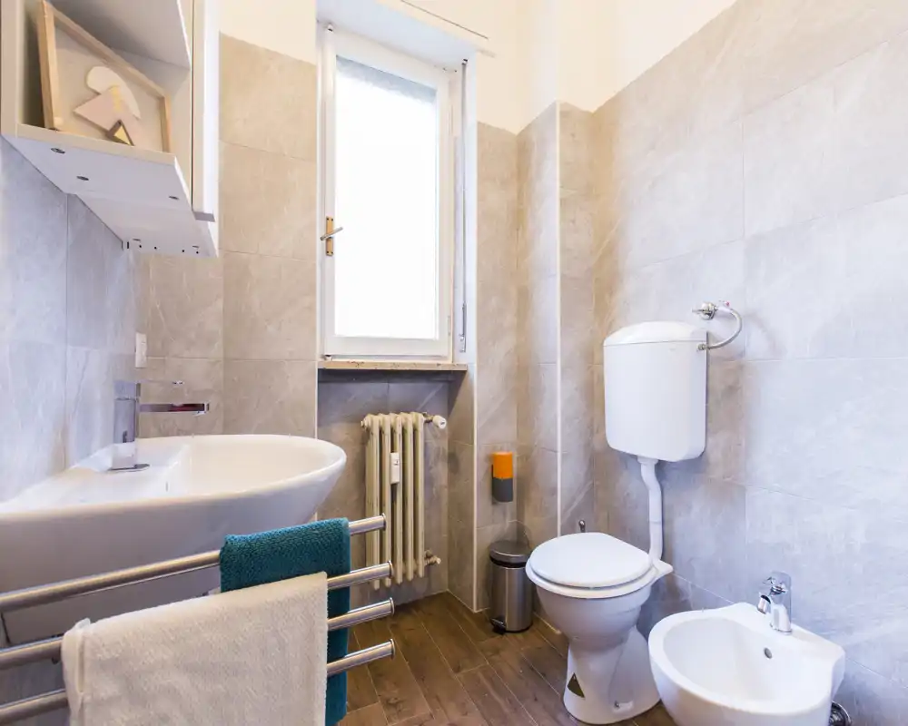 Bright, modern bathroom with a toilet, bidet, sink and window; light gray tiles and wood-effect floor create a clean, airy atmosphere.