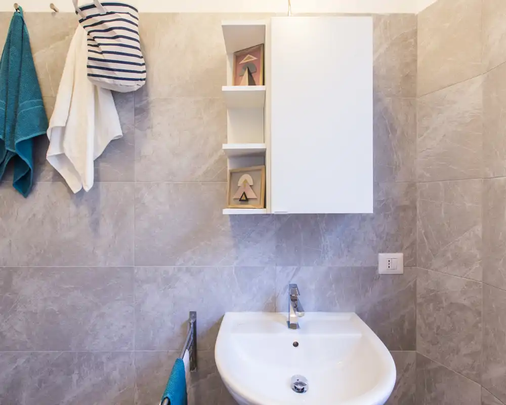 Small modern bathroom with a wall-mounted cabinet, sink and towel hooks; neutral gray stone tiles and bright, clean lighting.