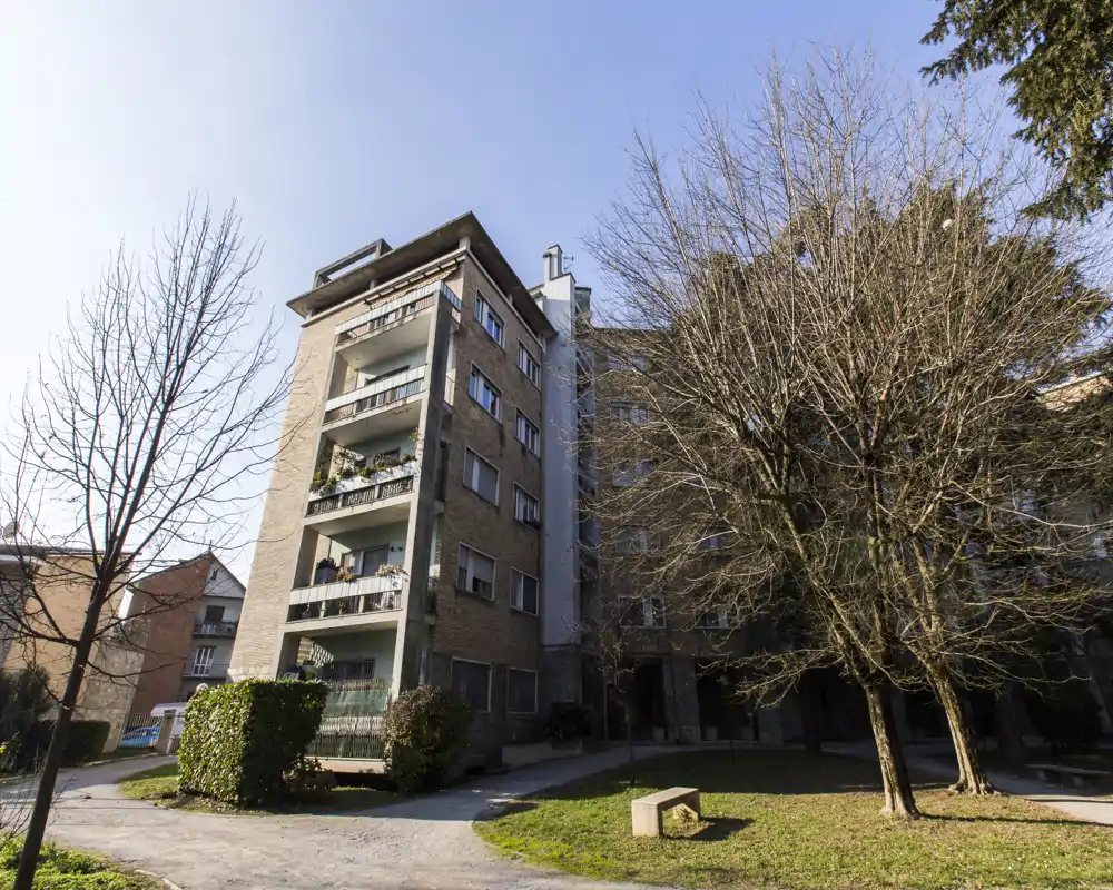 Exterior view of an apartment building and small courtyard with trees and benches, showing balconies and access paths — suitable for showcasing the building's façade and communal outdoor area.