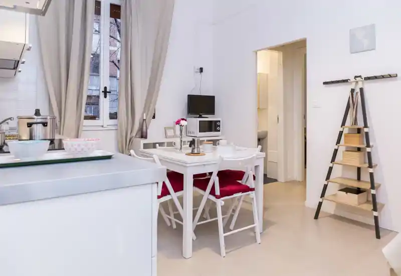 Bright, tidy dining corner in a compact kitchen area with a white table, four chairs with red cushions, and decorative shelving — ideal for small apartments.
