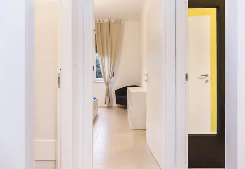 Bright, minimal bedroom viewed through an open doorway with a small bed edge, a curtain-covered window and a single armchair — simple, modern and well-lit.
