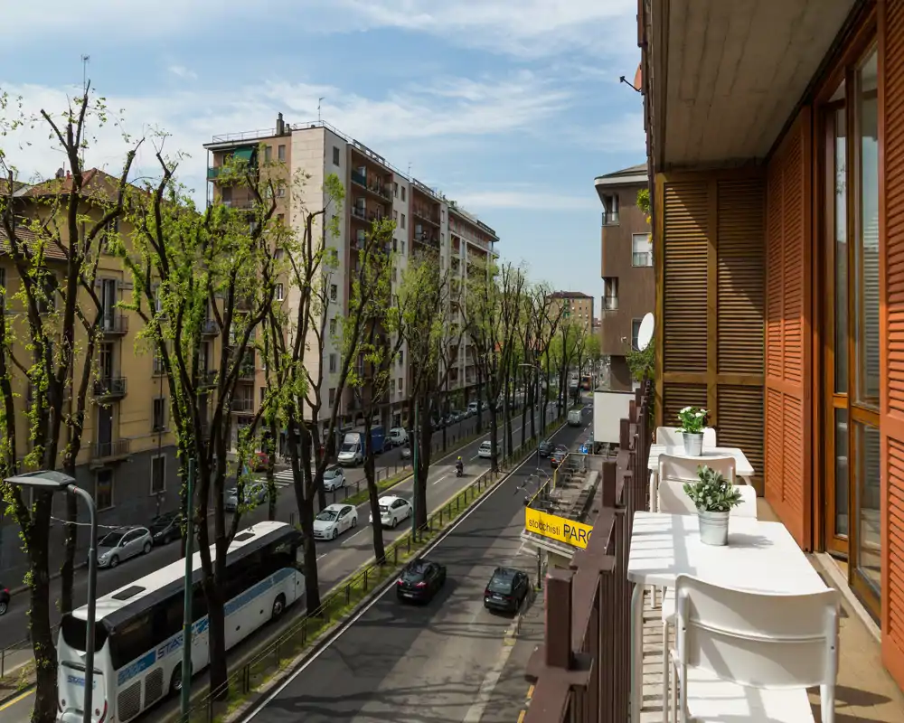 Balcón urbano soleado con vistas a una calle arbolada; pequeña mesa y sillas con macetas, ideal para el café de la mañana.