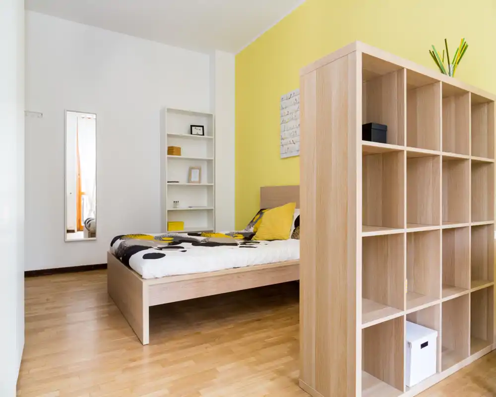 Dormitorio moderno y luminoso con cama de madera, acentos amarillos coordinados, estantería divisoria y decoración minimalista — perfecto para un alquiler compacto y con estilo.