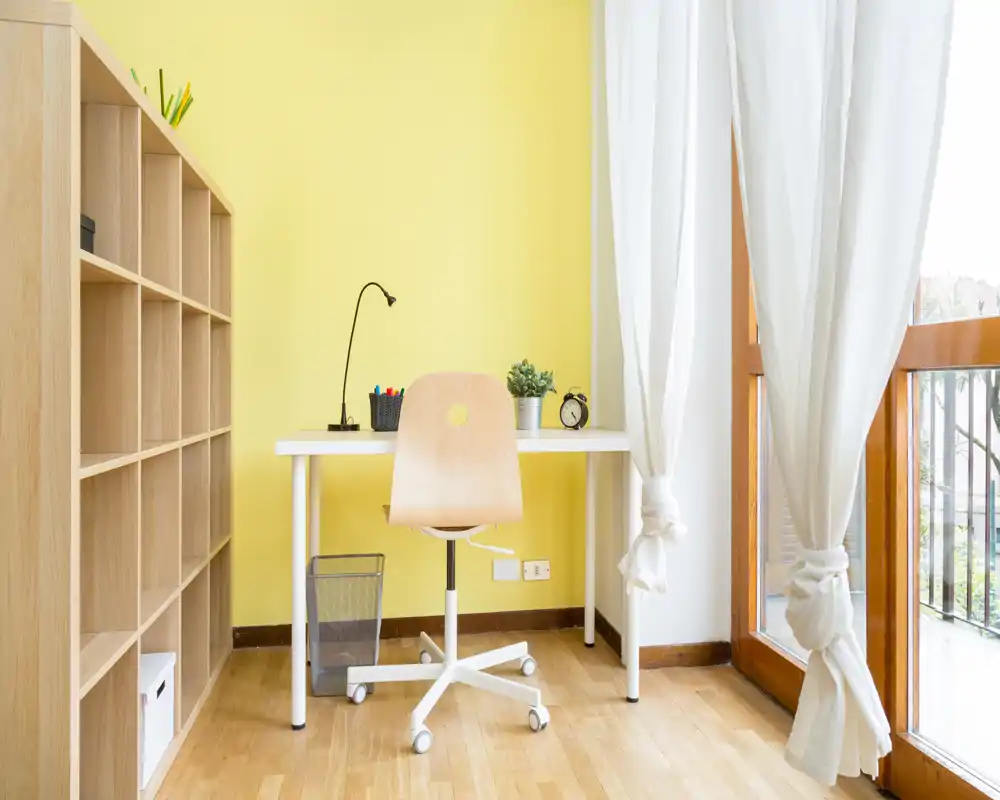 Rincón de oficina en casa luminoso y minimalista con un escritorio sencillo, silla con ruedas y estanterías junto a una gran ventana que aporta luz natural.