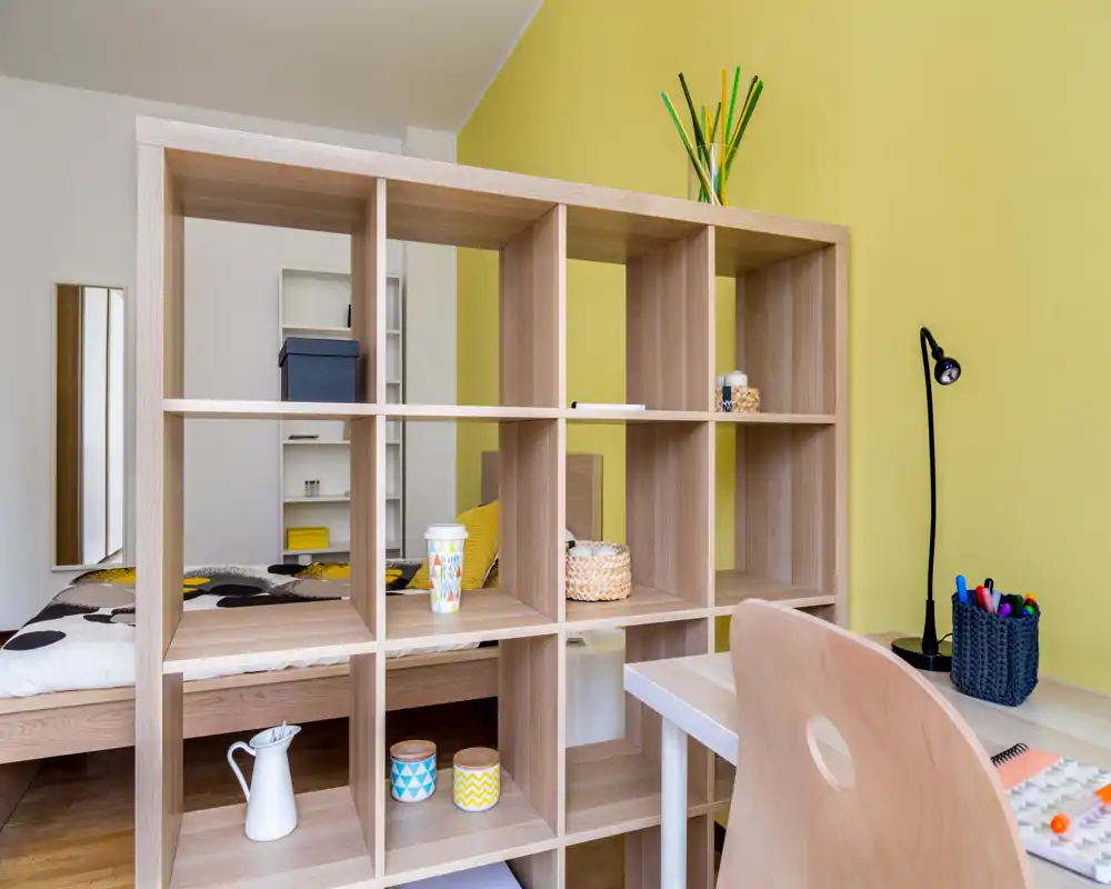 Dormitorio moderno y luminoso con un estante abierto de madera que separa la cama del espacio de trabajo. Luz natural, ropa de cama con estampado y cestas decorativas crean un ambiente acogedor y atractivo para los anuncios.