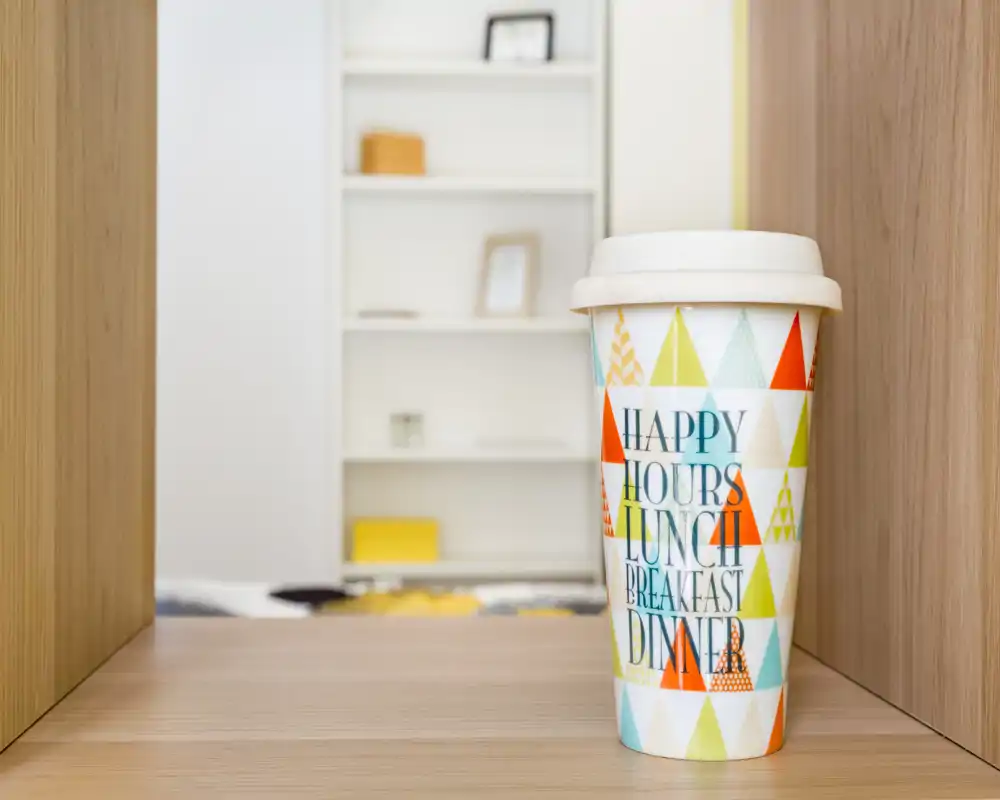 Primer plano de un vaso para llevar decorativo sobre una estantería de madera; al fondo se aprecia una estantería y textiles desenfocados, la función de la habitación no es identificable.