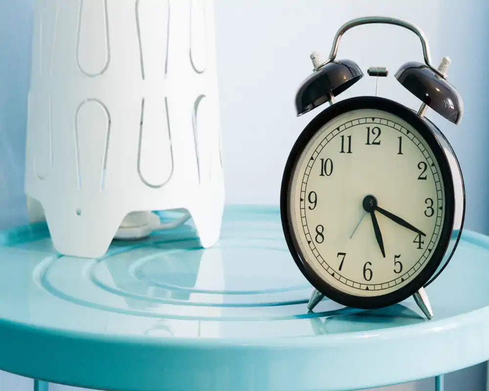 Primer plano de la superficie de una mesita con un despertador y parte de una lámpara decorativa; la imagen se centra en objetos más que en la habitación.