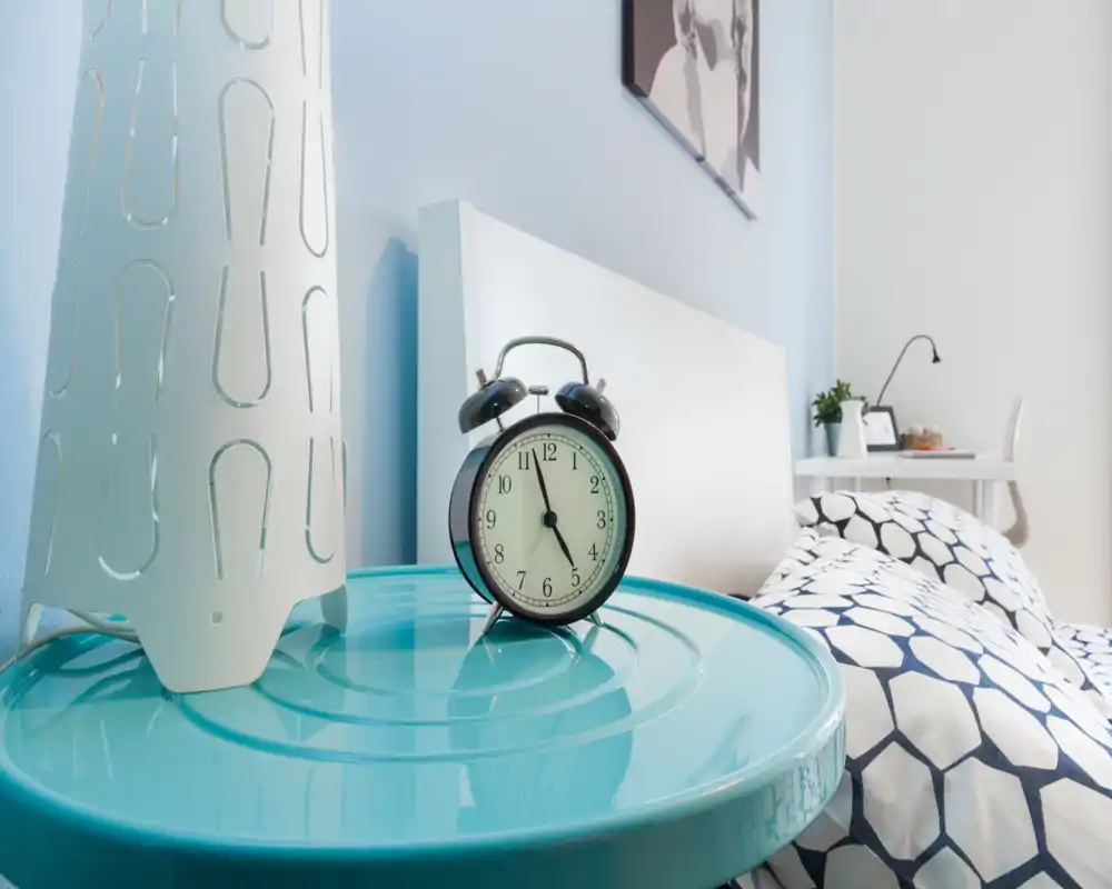 Rincón de dormitorio moderno y acogedor con ropa de cama estampada, cabecero blanco y mesita de noche con un reloj despertador retro y una lámpara decorativa. Toma luminosa y bien preparada, perfecta para un anuncio de alquiler que destaque los detalles.