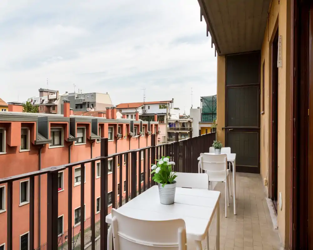 Balcón soleado con pequeñas mesas y sillas, macetas con plantas y vistas a los edificios cercanos — un espacio exterior compacto para el café por la mañana.