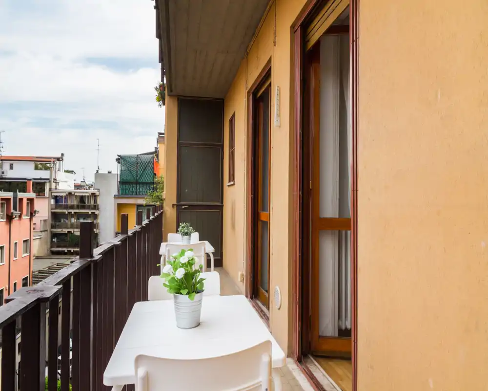 Balcón estrecho y soleado con pequeñas mesas blancas y plantas en maceta, con vistas a los edificios vecinos — ideal para el café de la mañana.