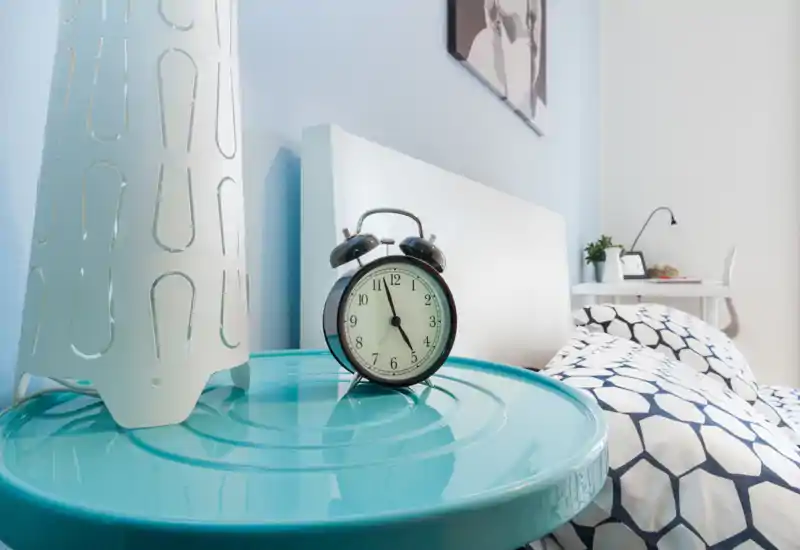 Rincón de dormitorio moderno y acogedor con ropa de cama estampada, cabecero blanco y mesita de noche con un reloj despertador retro y una lámpara decorativa. Toma luminosa y bien preparada, perfecta para un anuncio de alquiler que destaque los detalles.