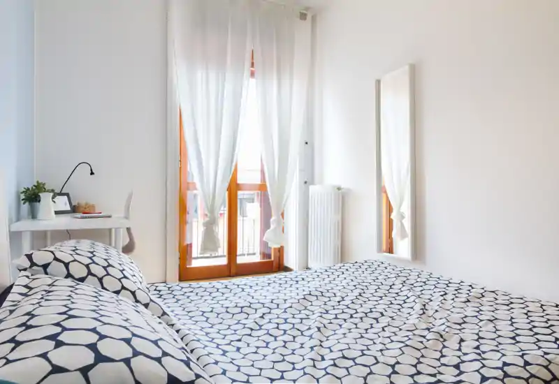 Dormitorio luminoso y minimalista con ropa de cama estampada, un pequeño escritorio junto a la ventana y una puerta al balcón que aporta mucha luz natural.