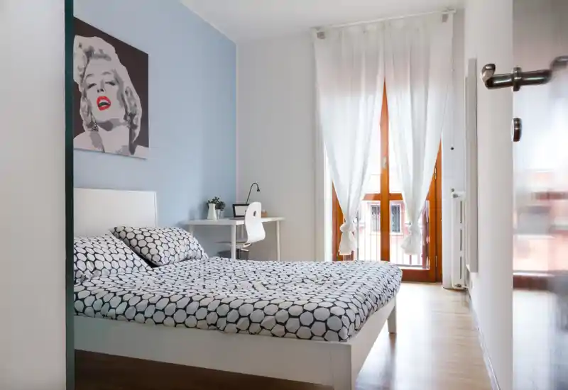 Dormitorio moderno y luminoso con cama doble grande, ropa de cama con estampado, pequeño escritorio y luz natural de la puerta a la terraza — limpio y con buen estilo.