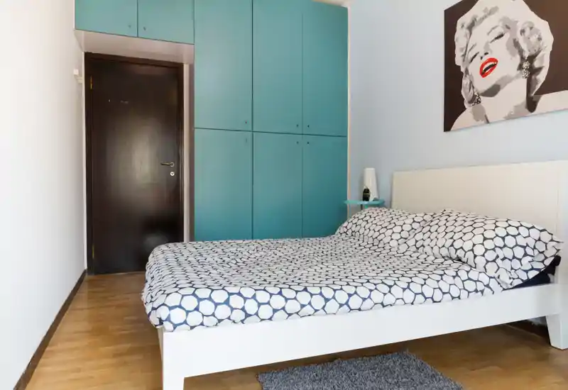 Dormitorio moderno y luminoso con cama doble estampada, armario empotrado turquesa y obra de arte en la pared. Suelo de madera y detalles en la mesita que hacen el espacio acogedor.