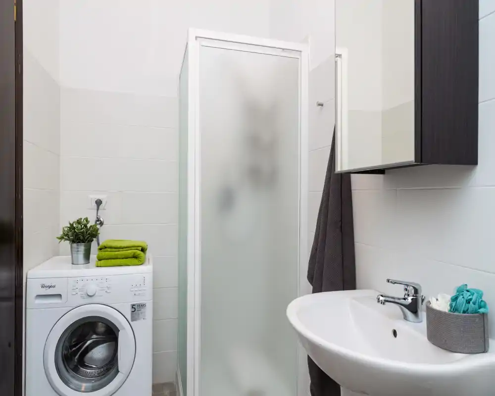 Baño compacto y moderno con cabina de ducha, lavabo de pared y lavadora junto a la ducha. Azulejos blancos, buena iluminación y distribución práctica, ideal para un piso pequeño.