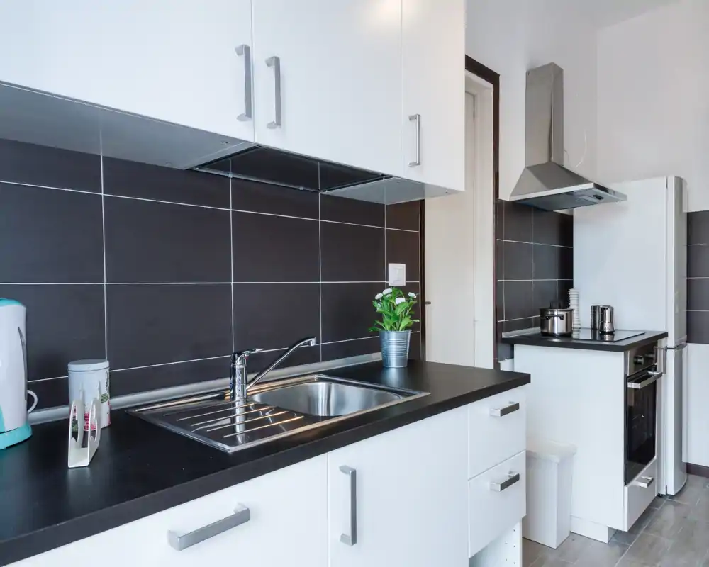 Cocina compacta y moderna con armarios blancos, fregadero de acero inoxidable, salpicadero oscuro de azulejos y horno integrado; ordenada y bien iluminada, perfecta para anuncios.