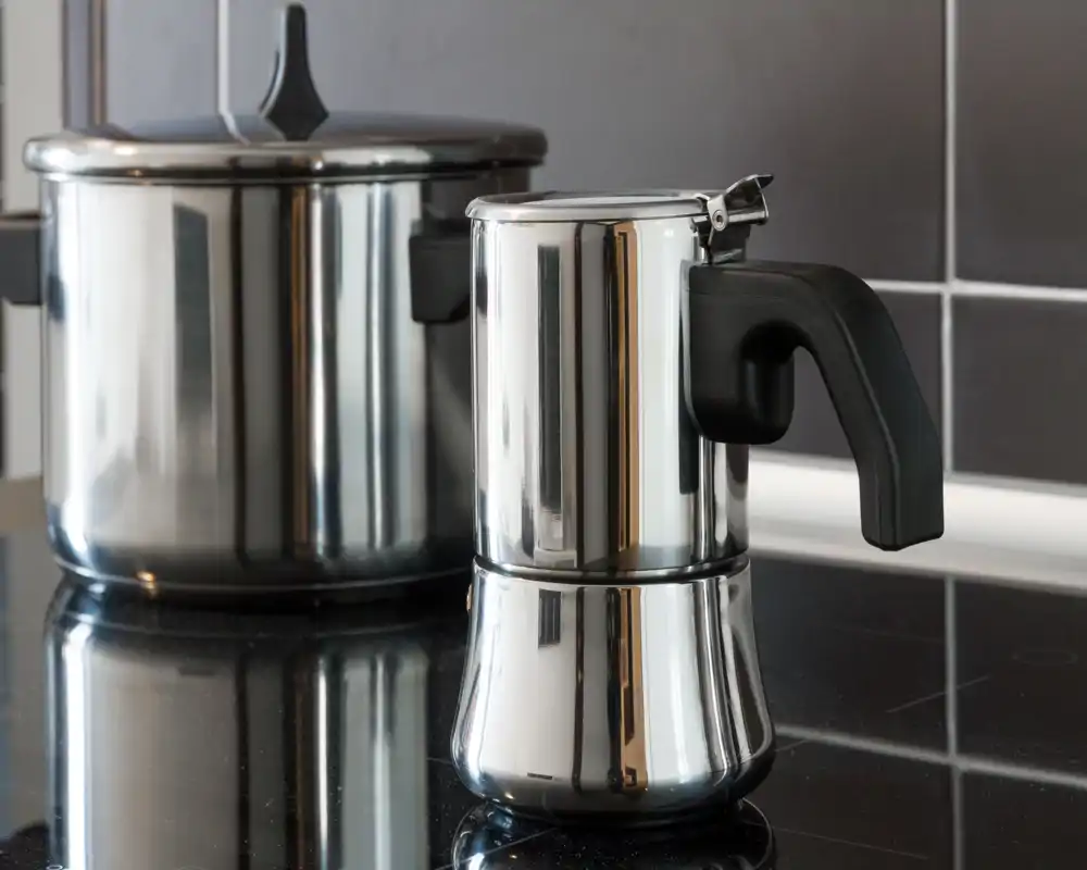 Primer plano de la cocina que muestra una cafetera moka de acero inoxidable y una olla grande sobre una placa vitrocerámica, con superficies reflectantes y azulejos modernos.