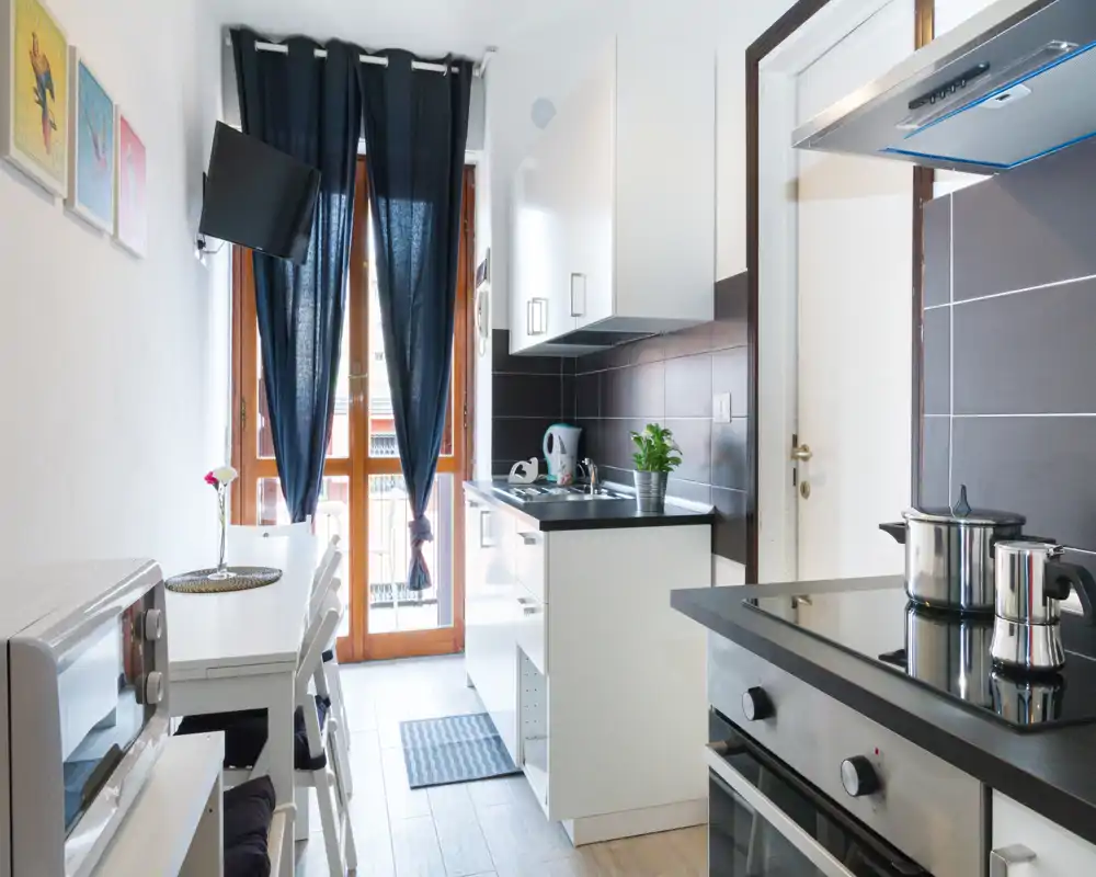 Cocina compacta y luminosa con acabados modernos en blanco y negro, horno y placa integrados, pequeña mesa comedor y luz natural desde la puerta de cristal.