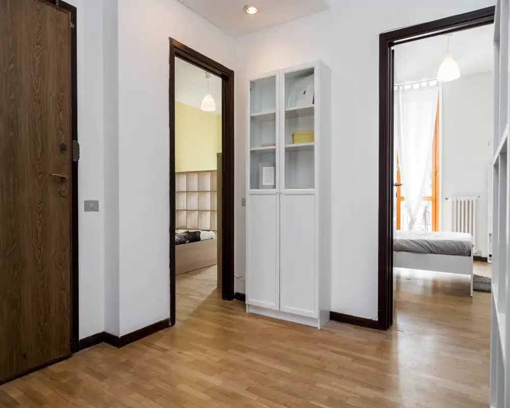 Recibidor luminoso y sencillo con suelo de madera y dos puertas que conducen a los dormitorios; un armario blanco alto apoyado en la pared.