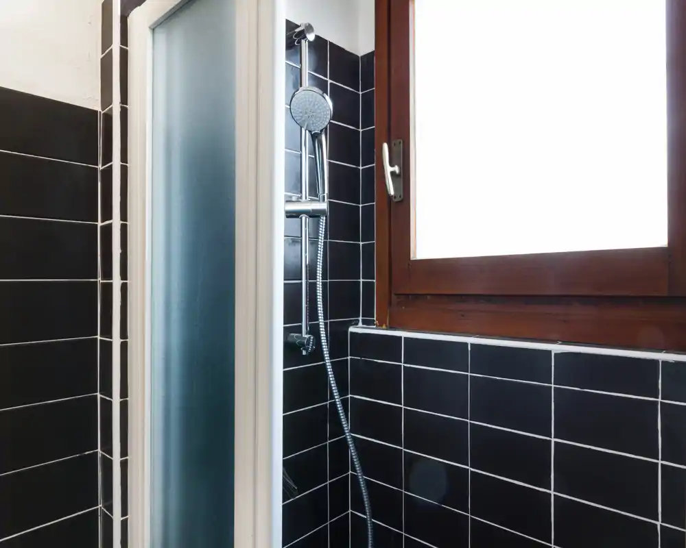 Ducha de baño compacta con azulejos negros, teleducha, puerta de ducha de vidrio esmerilado y ventana con marco de madera.