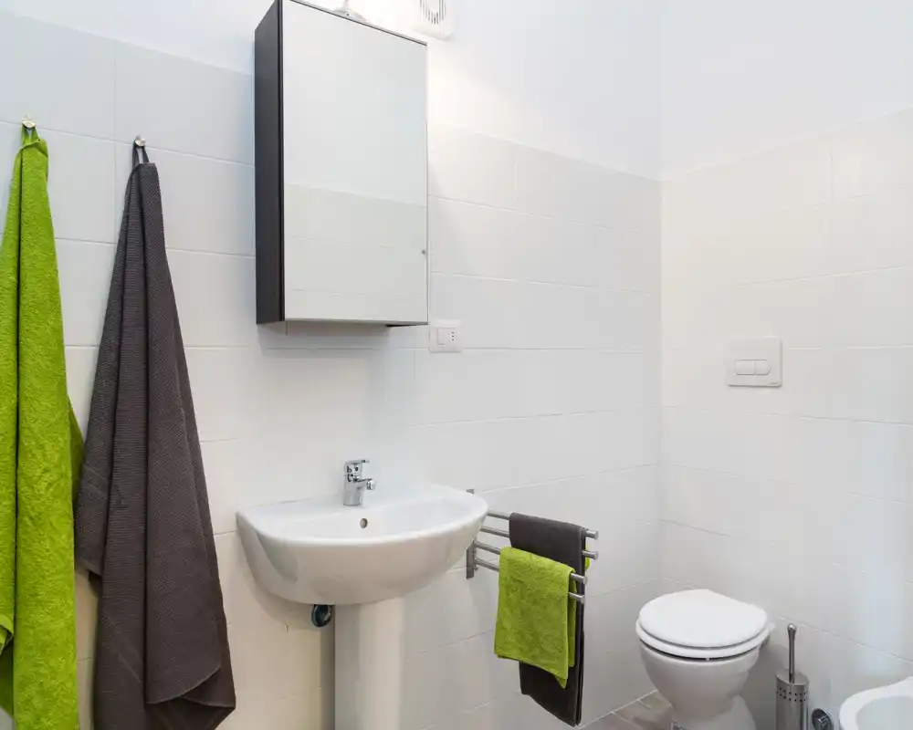 Baño limpio y bien iluminado con lavabo de pedestal, armario espejo, inodoro y accesorios cromados; azulejos blancos y toallas verdes/grises que aportan un toque fresco.