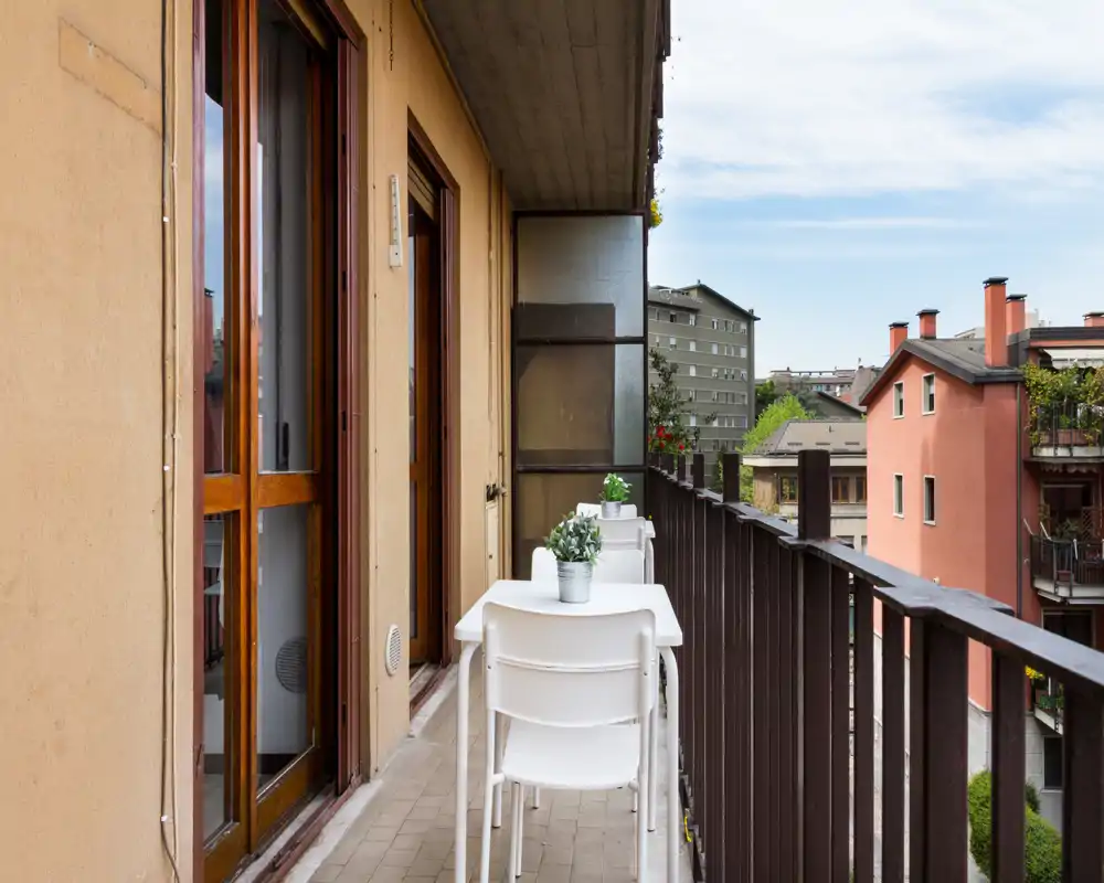 Balcón estrecho y soleado con dos sillas blancas y una mesita con plantas en maceta, con vistas a los edificios cercanos — ideal para el café de la mañana.