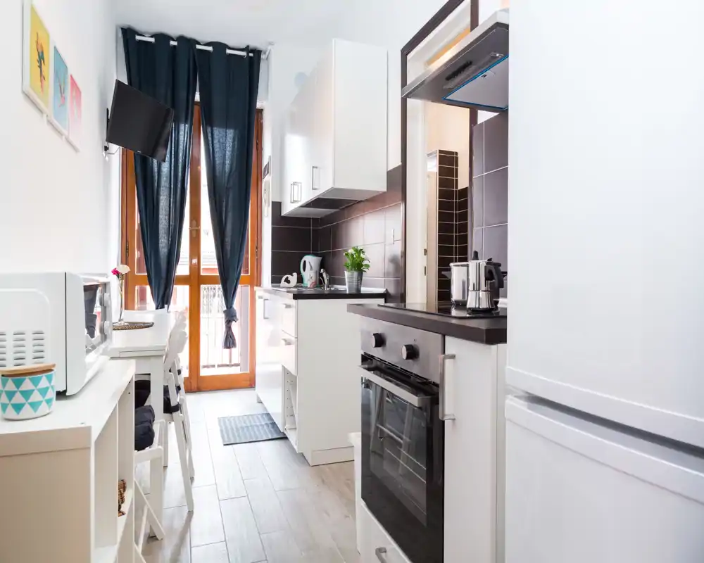 Cocina moderna tipo galería, luminosa, con armarios blancos, horno empotrado, mesa pequeña y puerta hacia el balcón que aporta luz natural.