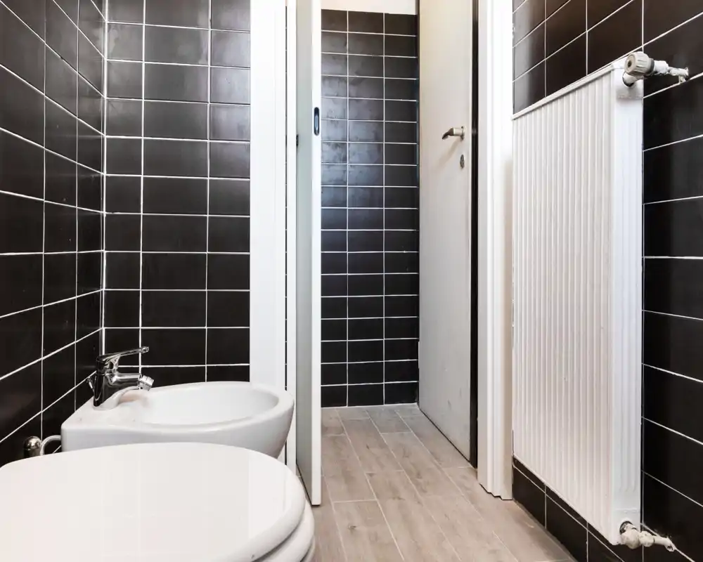 Baño compacto y moderno con paredes de azulejos negros, bidé y váter visibles y un radiador vertical; suelo efecto madera para un aspecto limpio y contemporáneo.