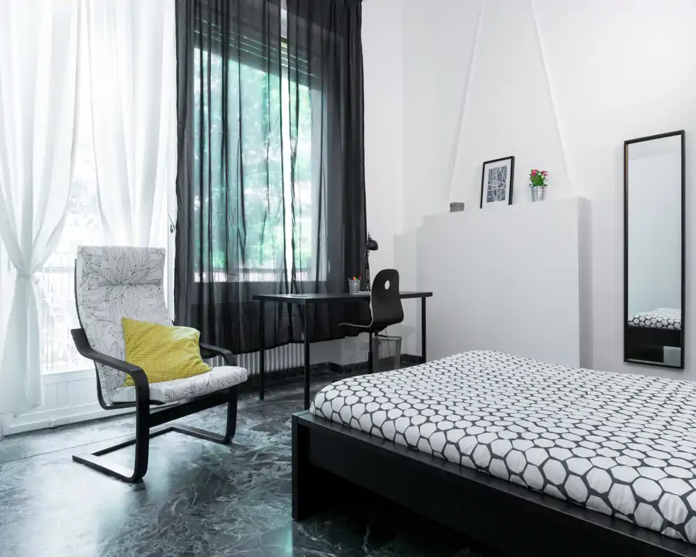 Dormitorio moderno y luminoso con ropa de cama estampada, un sillón de lectura con cojín amarillo, escritorio junto a la ventana y cortinas negras translúcidas que dejan pasar la luz natural.