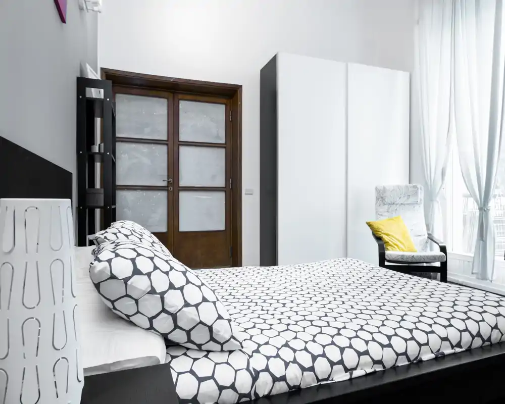Dormitorio moderno y luminoso con ropa de cama estampada, un gran armario y un sillón junto a la ventana. La luz natural inunda la estancia, creando un ambiente limpio y acogedor perfecto para anuncios.