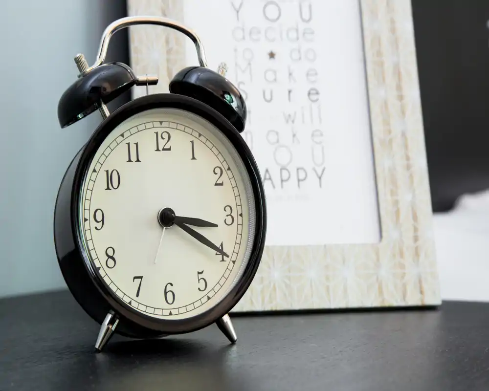 Primer plano de un despertador sobre la mesita de noche con un marco en segundo plano, que sugiere un ambiente de dormitorio y una decoración acogedora.