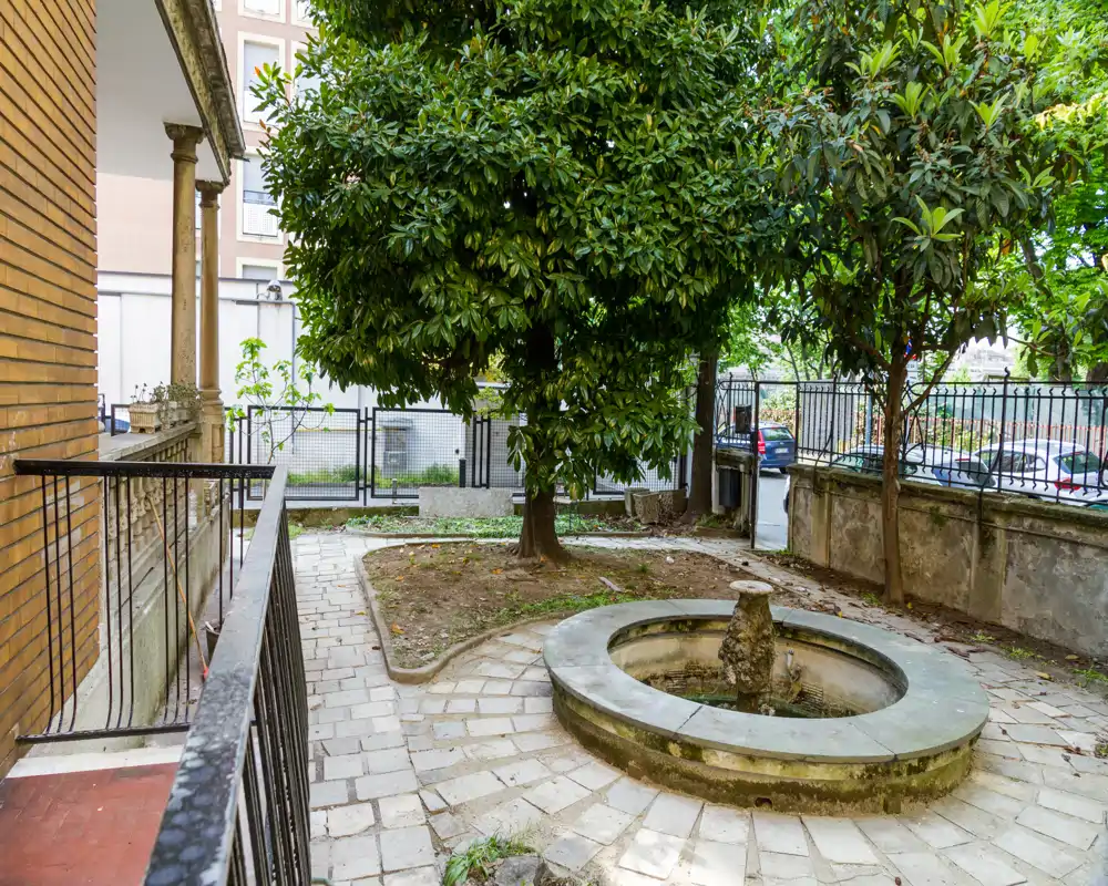 Pequeño patio-jardín con una fuente circular de piedra, camino pavimentado y árboles maduros que proporcionan sombra — un rincón exterior tranquilo.